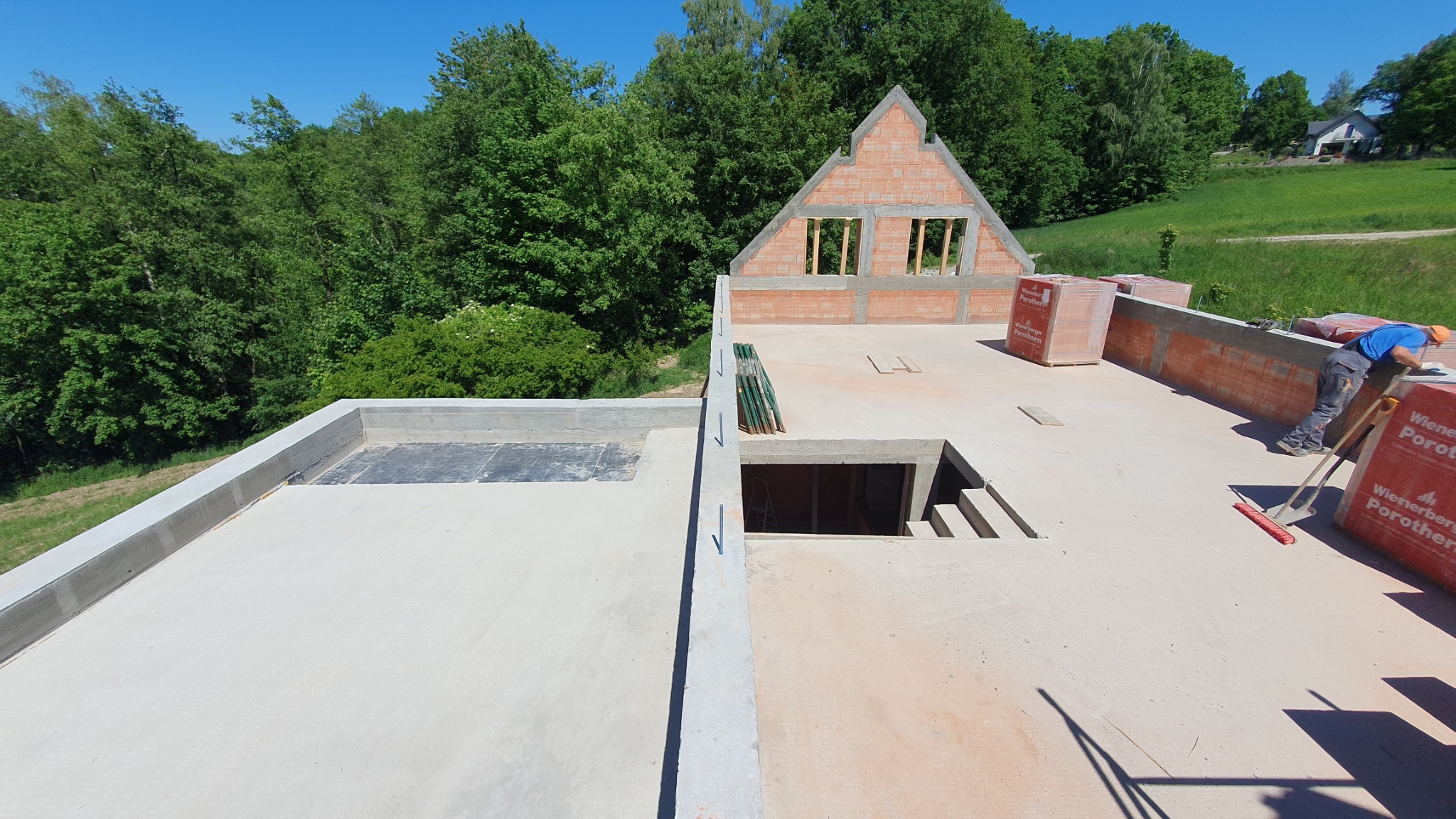 Wznoszenie ścian domu z pustaków ceramicznych na betonowej płycie fundamentowej z widocznym otworem na schody; w tle zielone drzewa i częściowo zabudowany teren.