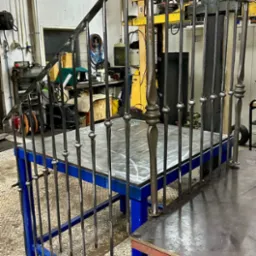 Surowa, metalowa konstrukcja schodów z balustradą, w warsztacie ślusarskim, widoczne elementy dekoracyjne na słupkach, spawana na niebieskim stole warsztatowym.