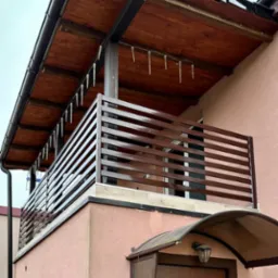 Brązowa balustrada balkonowa z poziomych profili na tle różowej elewacji domu, ozdobiona lampkami w kształcie sopli lodu.