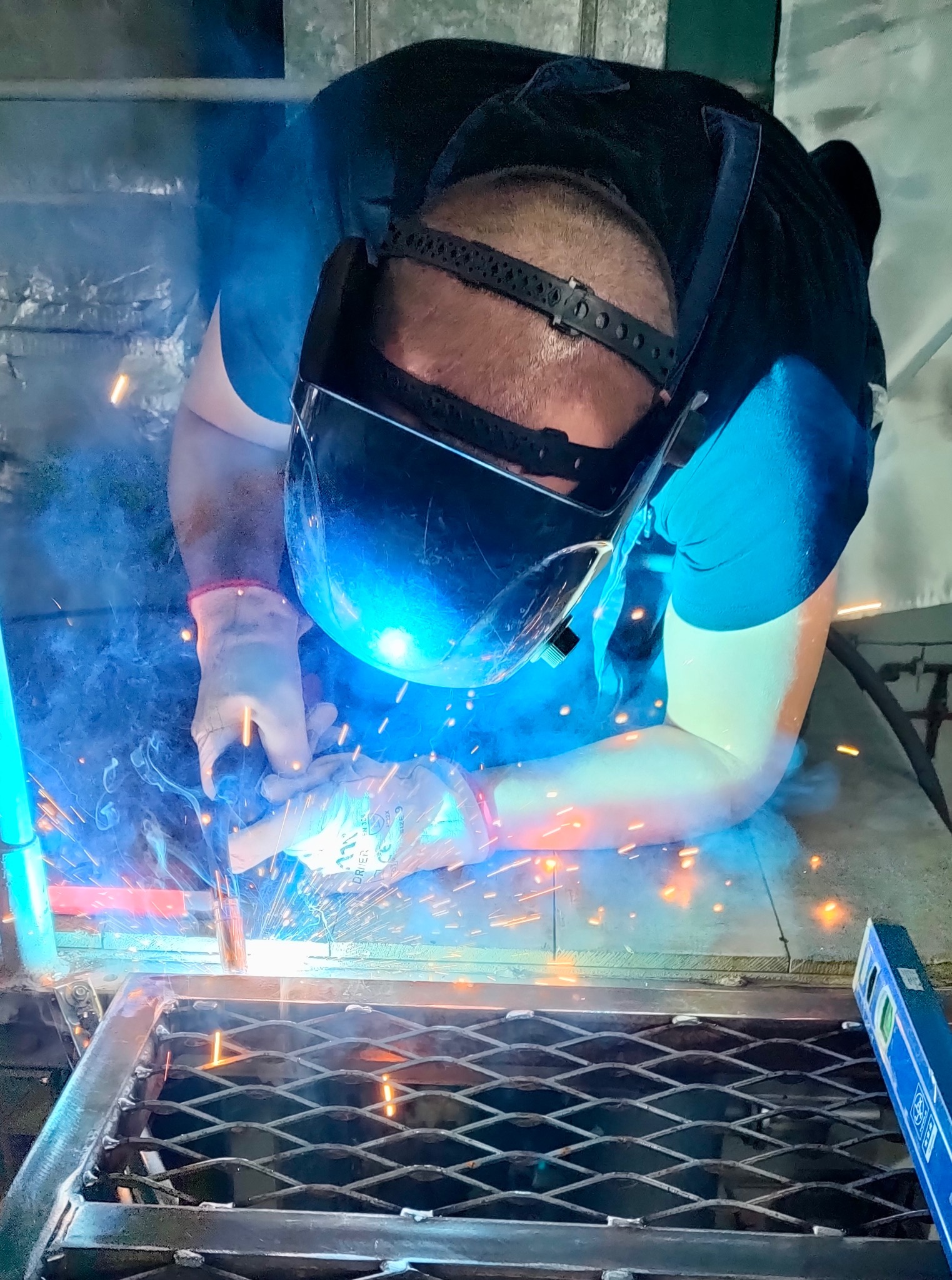 Spawanie metalowej konstrukcji kratowej; spawacz w masce ochronnej, iskry i dym w warsztacie, poziomica obok