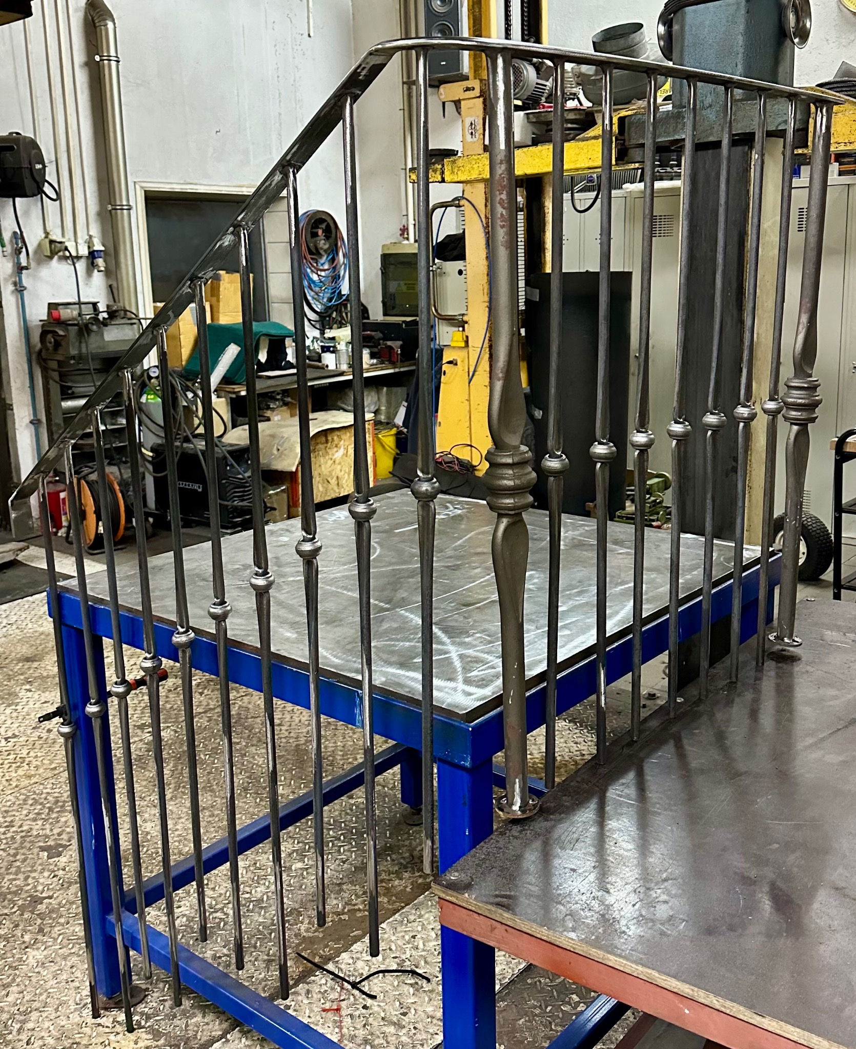 Surowa, metalowa konstrukcja schodów z balustradą, w warsztacie ślusarskim, widoczne elementy dekoracyjne na słupkach, spawana na niebieskim stole warsztatowym.