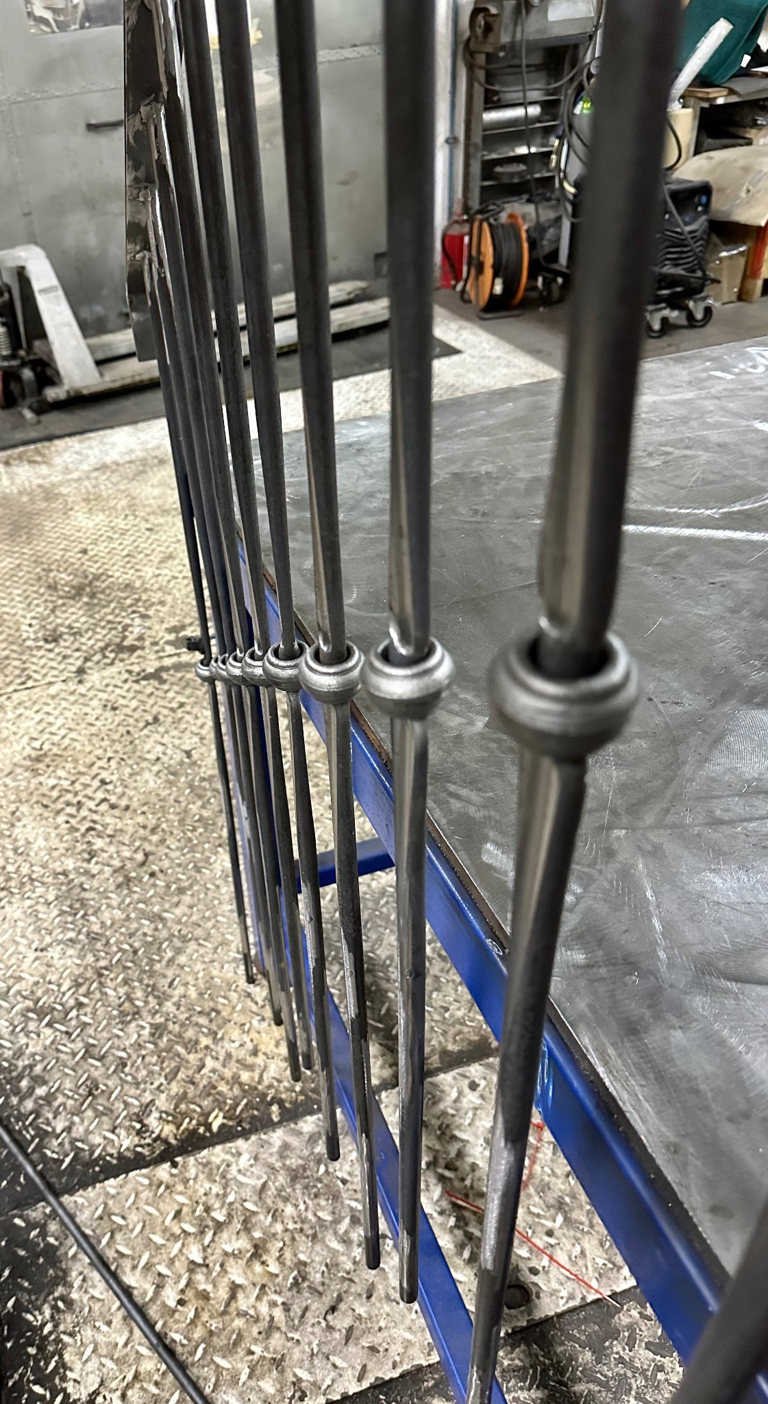 Metalowe pręty z ozdobnymi kulistymi elementami, prawdopodobnie fragment balustrady lub ogrodzenia w trakcie produkcji w warsztacie ślusarskim, widoczne narzędzia i wyposażenie warsztatu w tle.