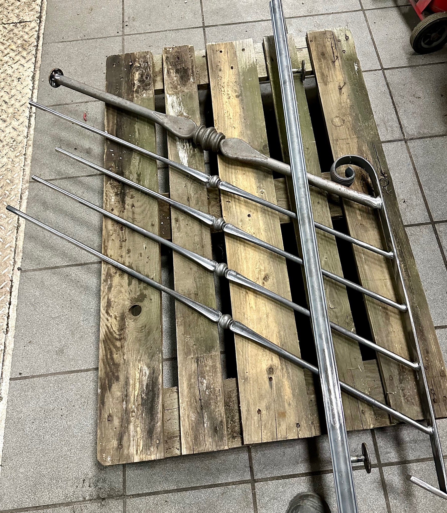 Elementy metalowe balustrady, w tym ozdobne pręty i gięta poręcz, ułożone na drewnianej palecie w warsztacie.
