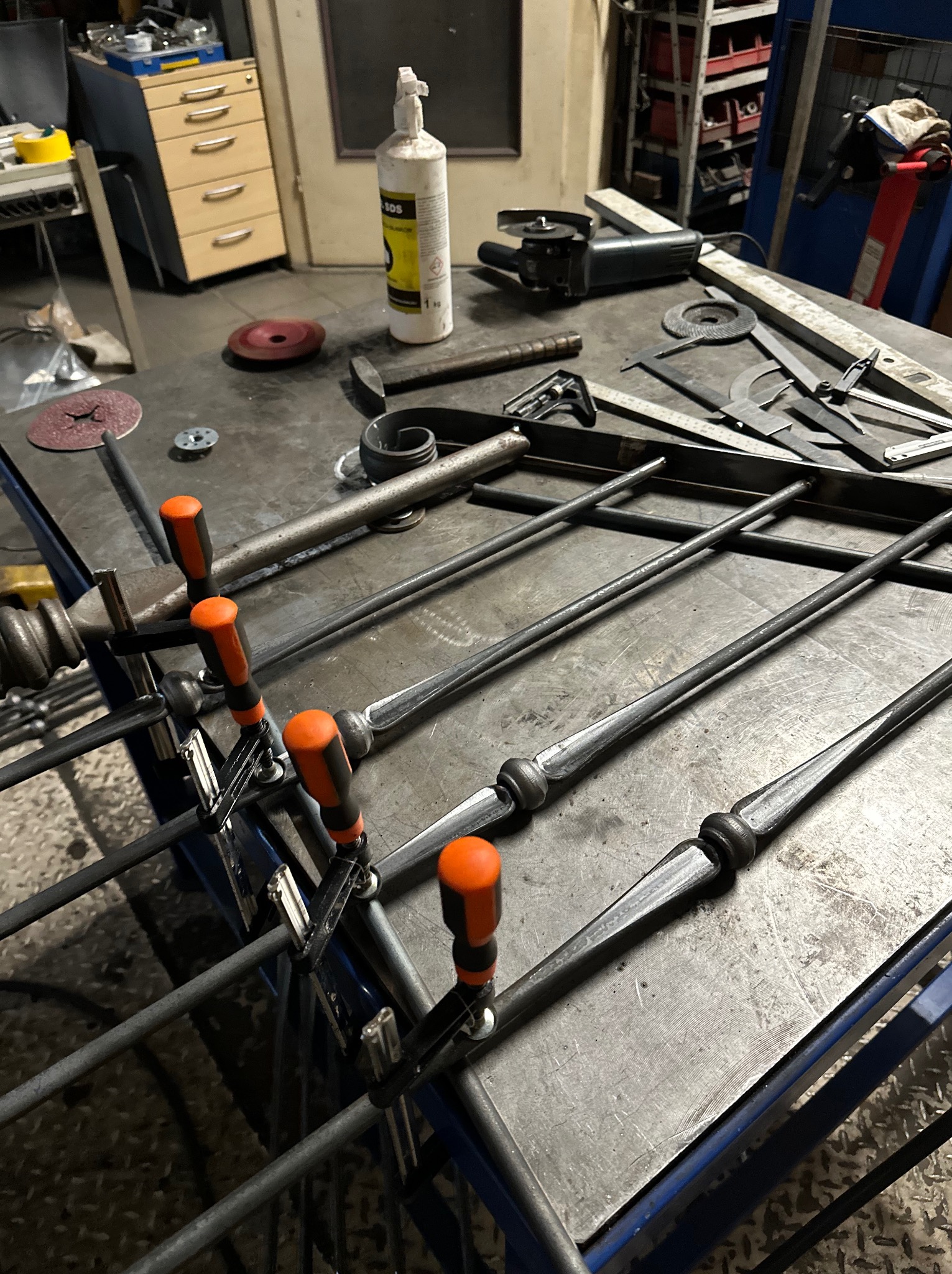 Metalowe pręty z ozdobnymi elementami, mocowane do ramy za pomocą zacisków stolarskich na stole warsztatowym, w tle narzędzia ślusarskie i szafka z szufladami.