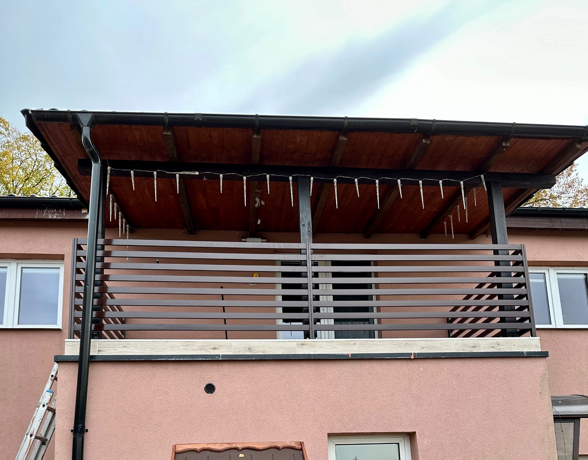 Zadaszony balkon z poziomą balustradą z brązowych listew, ozdobiony sznurem lampek w kształcie sopli, widok z dołu na tle pochmurnego nieba.