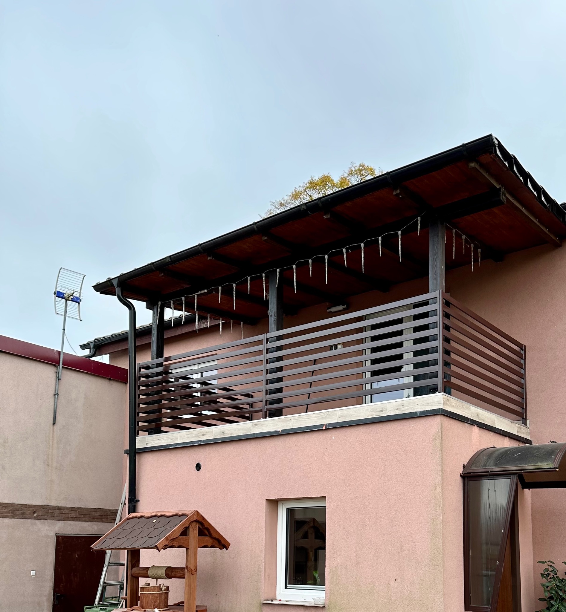 Balkon z brązową, poziomą balustradą, drewnianym zadaszeniem z imitacją sopli lodu, studnią zadaszoną gontem i drabiną opartą o ścianę budynku.