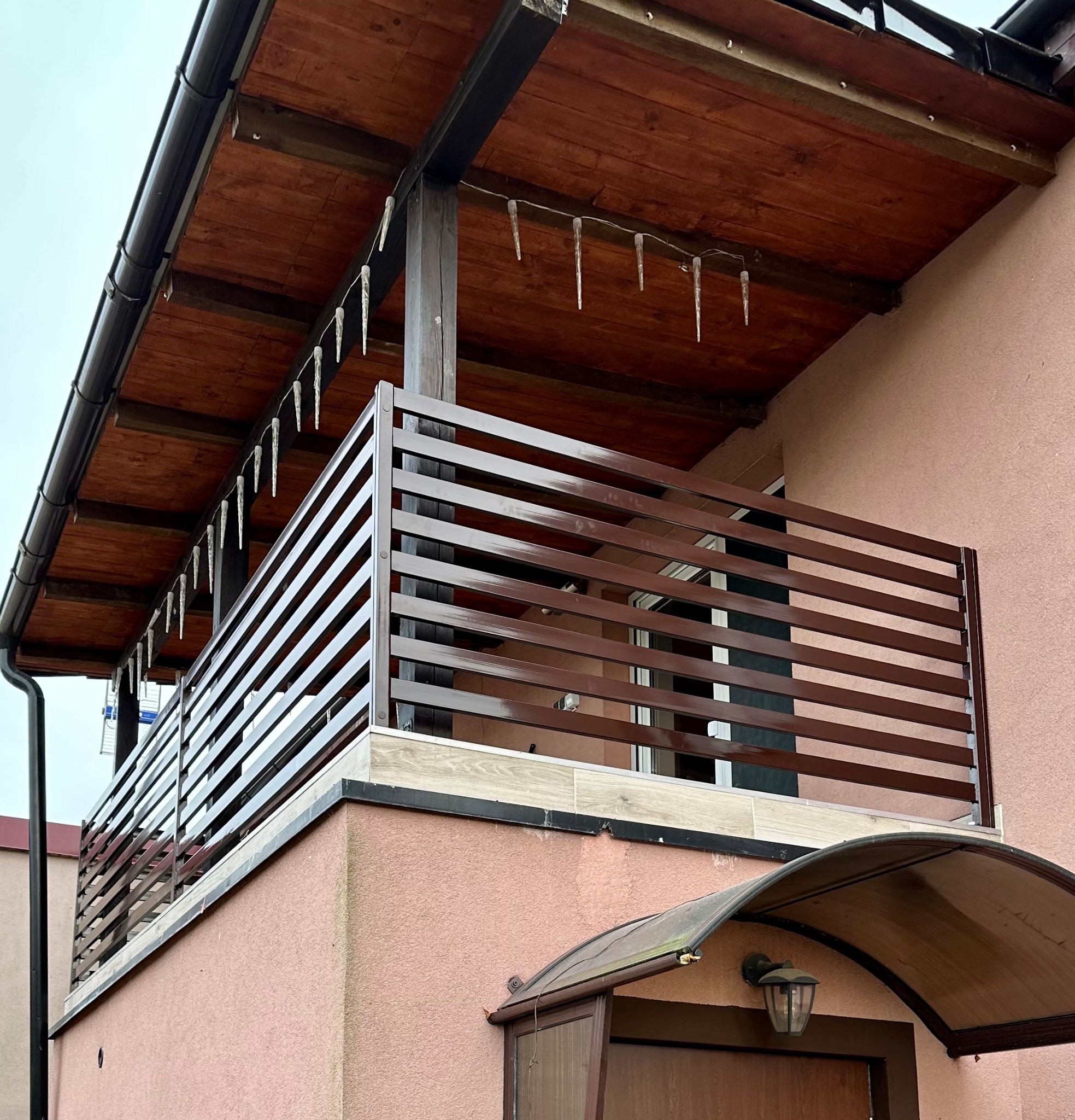 Brązowa balustrada balkonowa z poziomych profili na tle różowej elewacji domu, ozdobiona lampkami w kształcie sopli lodu.