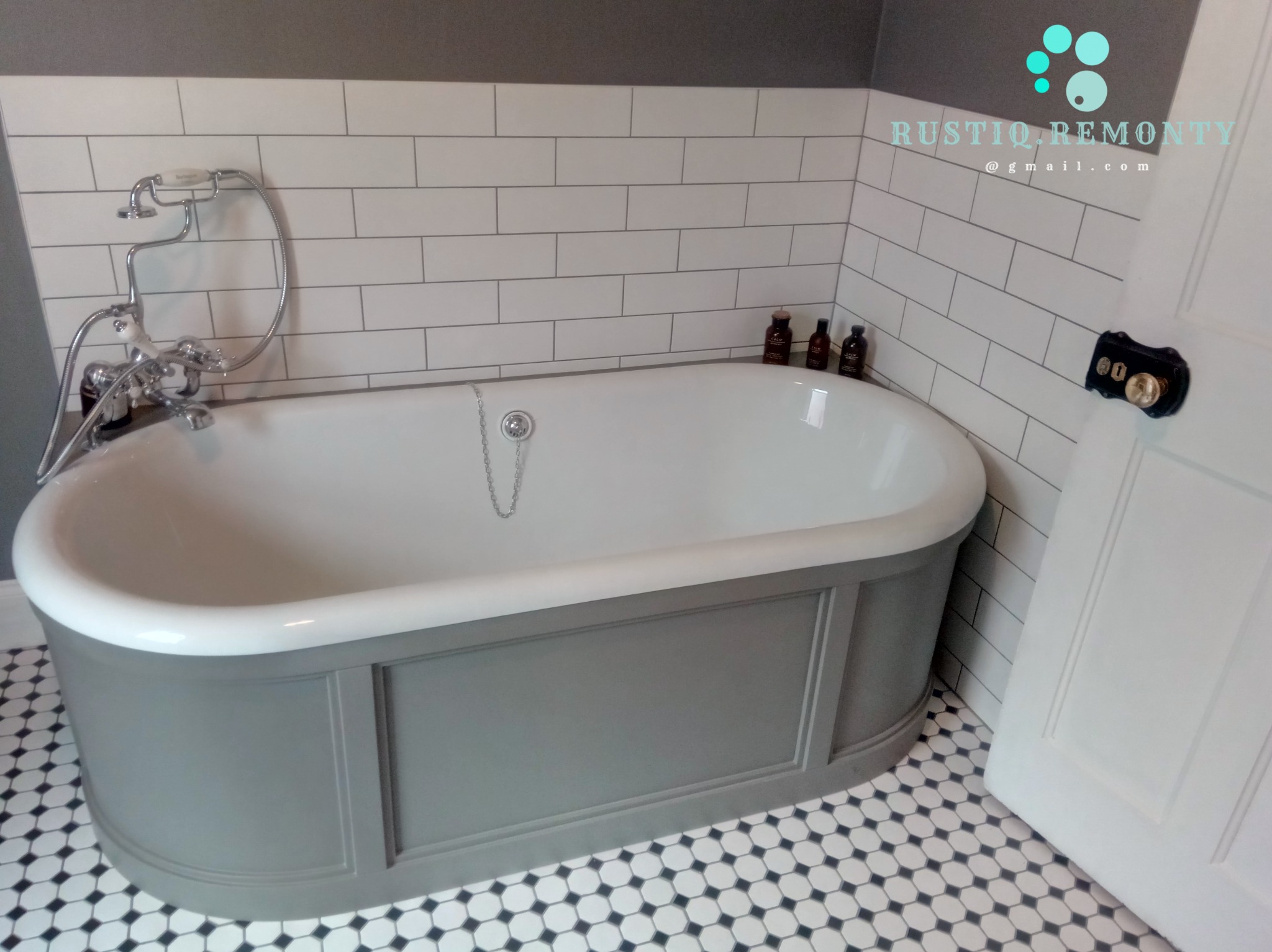 Wolnostojąca wanna na tle białych, prostokątnych płytek i mozaiki podłogowej w czarno-białe kropki. Szare panele dekoracyjne obudowują wannę. Na wannie stoją trzy brązowe buteleczki. Widoczny...