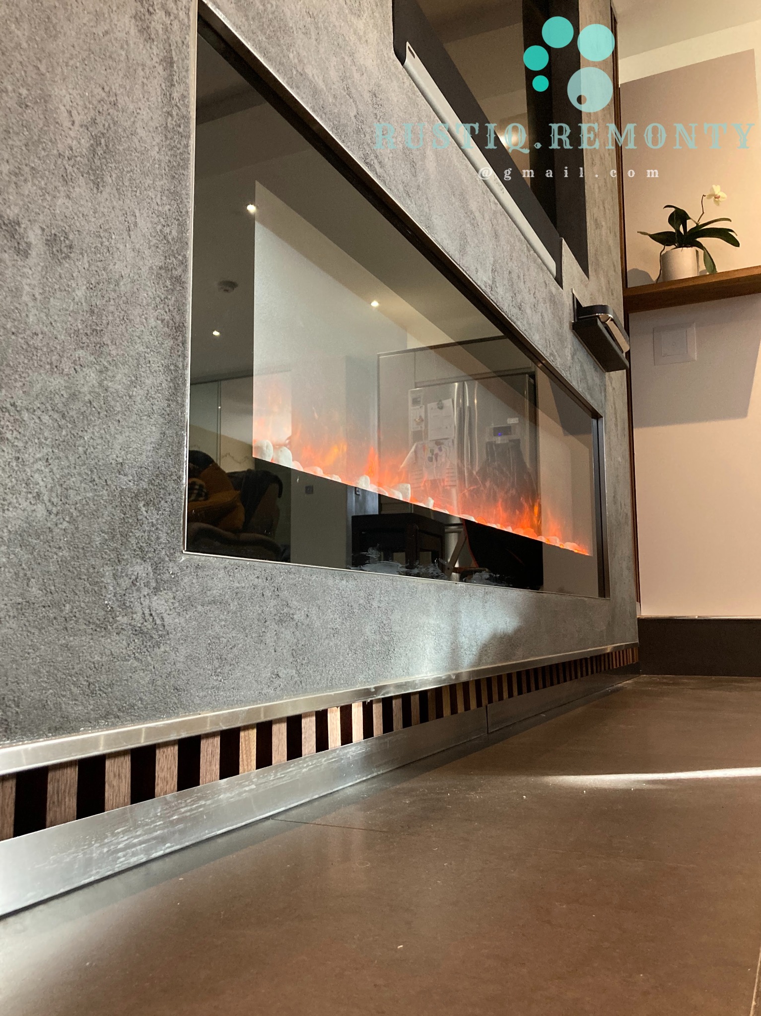 Nowoczesny kominek elektryczny w szarej obudowie z efektem płomieni, widoczne logo firmy i doniczka z kwiatem na półce.