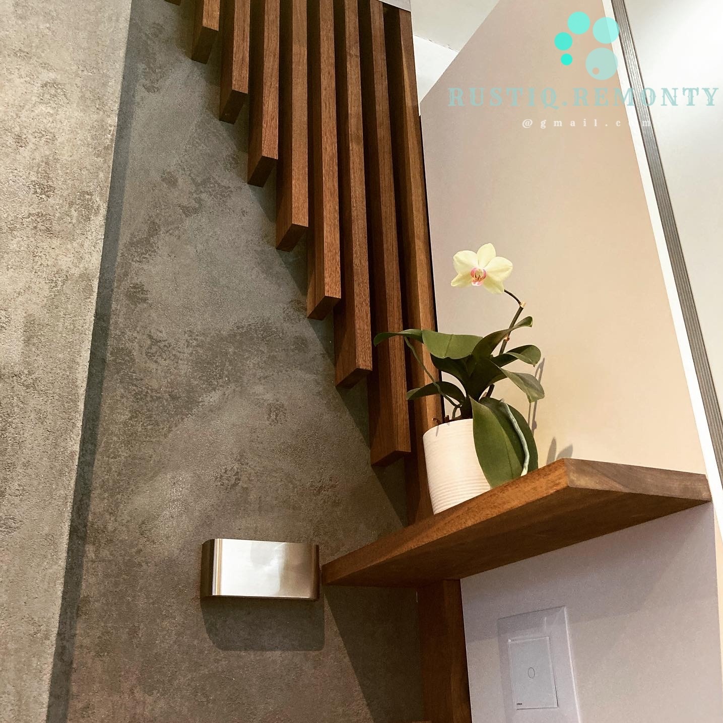 Fragment ściany z dekoracyjnym betonem, drewnianą lamelą w spadającym układzie, półką z orchideą i metalowym uchwytem na papier toaletowy.