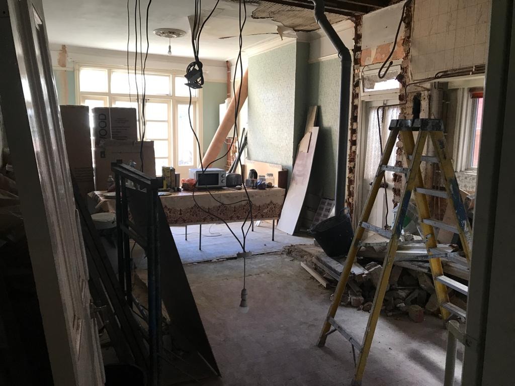 Pokój w trakcie demontażu: odsłonięte ściany, wiszące przewody, drabina oparta o ścianę, gruz na podłodze, zabezpieczone meble w tle.
