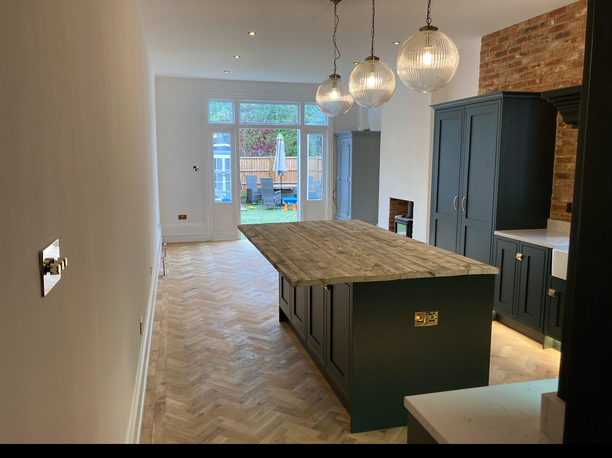 Nowoczesna kuchnia z ciemnozielonymi szafkami, drewnianym blatem wyspy i parkietem w jodełkę, widok na ogród przez przeszklone drzwi.