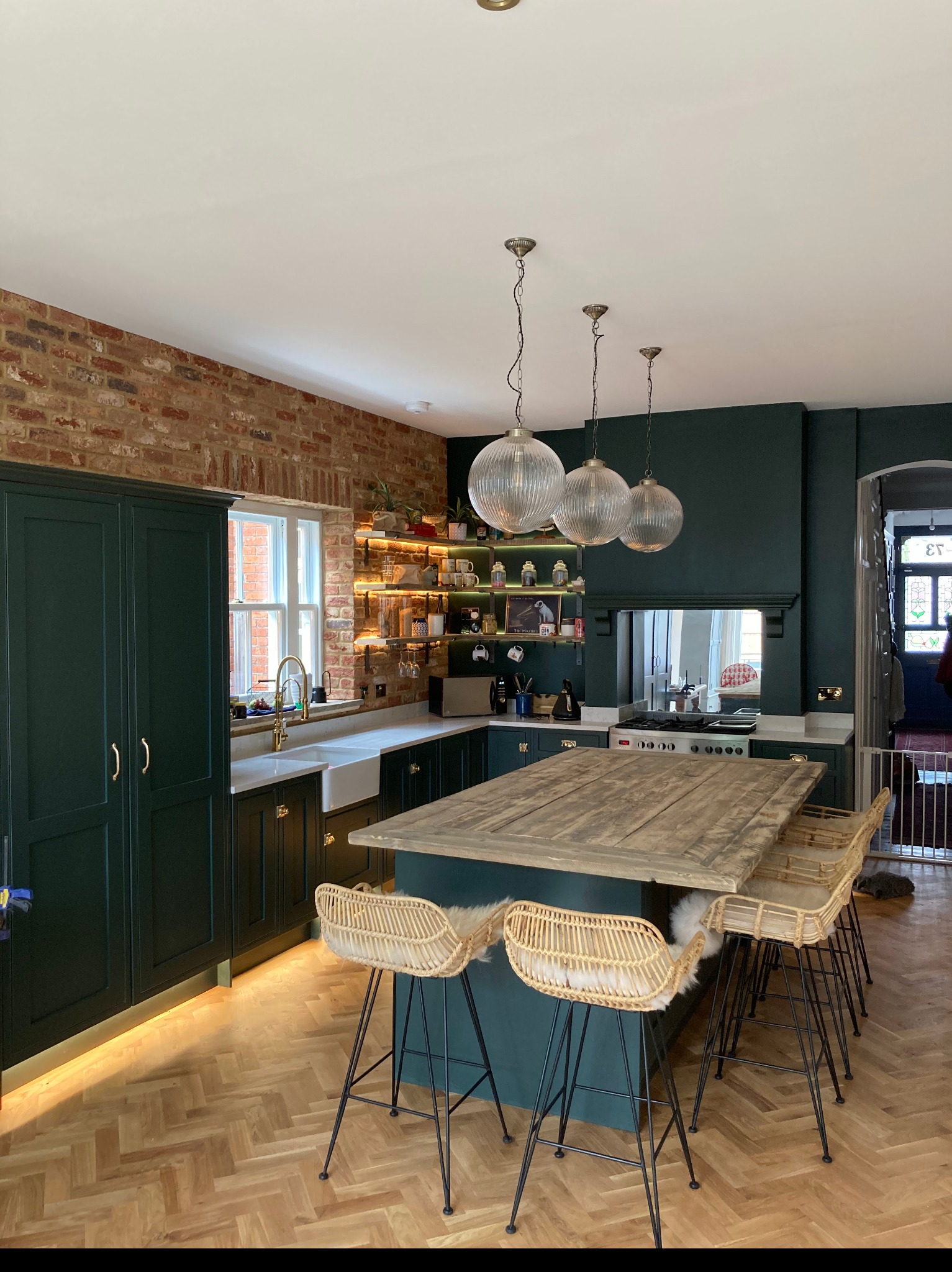 Kuchenne wnętrze w odcieniach butelkowej zieleni, z ceglaną ścianą, drewnianym parkietem w jodełkę i wyspą z blatem z postarzanego drewna; trzy szklane lampy wiszące nad wyspą.