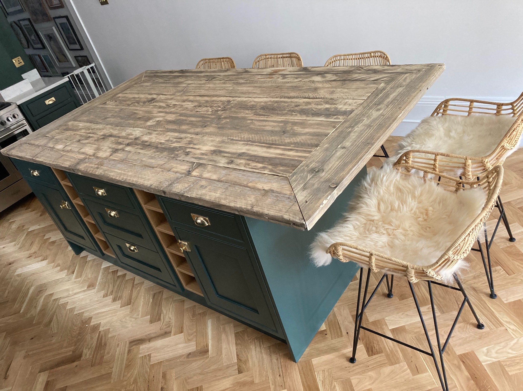 Wyspa kuchenna z blatem z postarzanego drewna i ciemnozielonymi szafkami, uzupełniona o rattanowe krzesła z futrzanymi poduszkami, na podłodze parkiet w jodełkę.