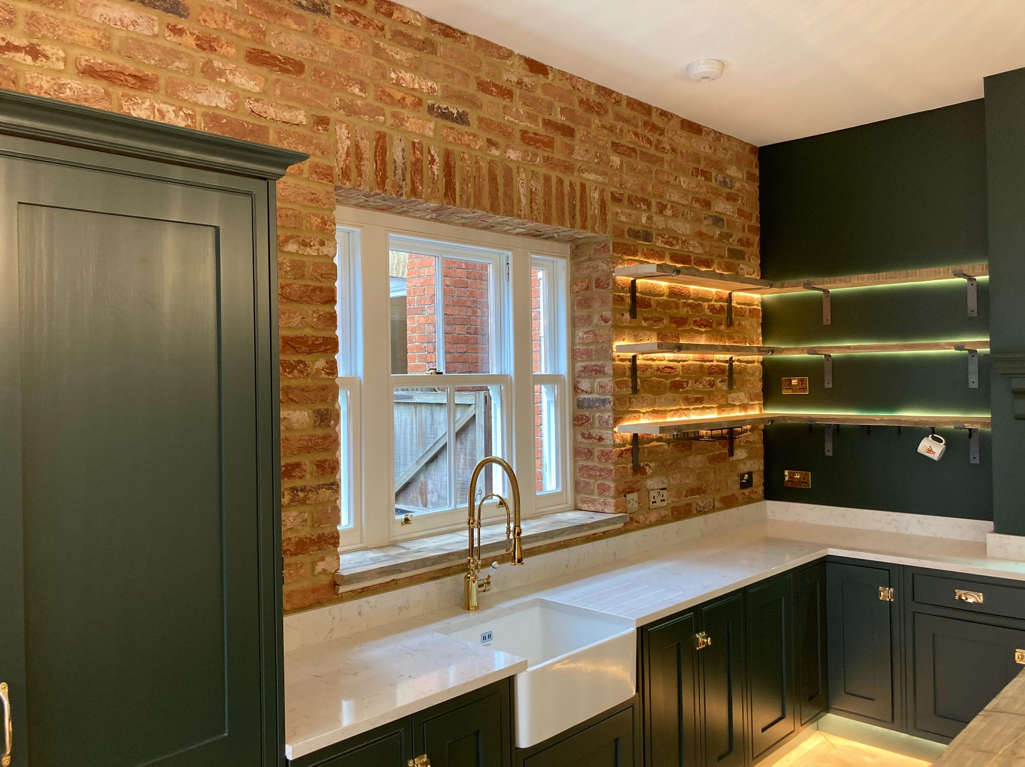 Kuchnia z ciemnozielonymi szafkami, białym blatem, ceglaną ścianą i złotą armaturą, oświetlone półki z kubkiem wiszącym na jednej z nich.