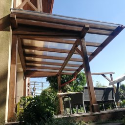 Woodesigne - Zadaszenie tarasu z brązowego drewna, z przezroczystym dachem, widok z dołu na tle błękitnego nieba, z widocznymi meblami ogrodowymi i zielenią.