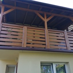 Woodesigne - Zadaszony balkon z drewnianą balustradą z poziomych desek, widok z dołu, doniczkowe rośliny na balkonie, brązowe rynny.