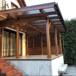 Woodesigne - Nowoczesna, drewniana konstrukcja z zadaszeniem, prawdopodobnie taras lub weranda, z ażurowymi ściankami i schodami z płytek, na tle zieleni.