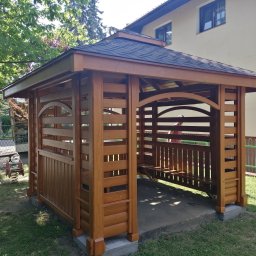 Woodesigne - Drewniana altana ogrodowa z dachem krytym gontem bitumicznym, z ławką wewnątrz, na betonowej podstawie, na tle zieleni i budynku. Widoczne detale wykończenia drewna.