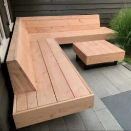 Drewniana, narożna ławka z niskim stolikiem na tarasie z szarych płyt. Prosta konstrukcja z jasnego drewna, w tle ściana budynku i wysoka trawa.