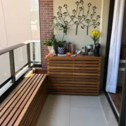 Funkcjonalny balkon z drewnianą zabudową, siedziskiem i dekoracjami. Wyjątkowy detal: metalowa ozdoba ścienna w kształcie drzewa. Jasne płytki podłogowe.