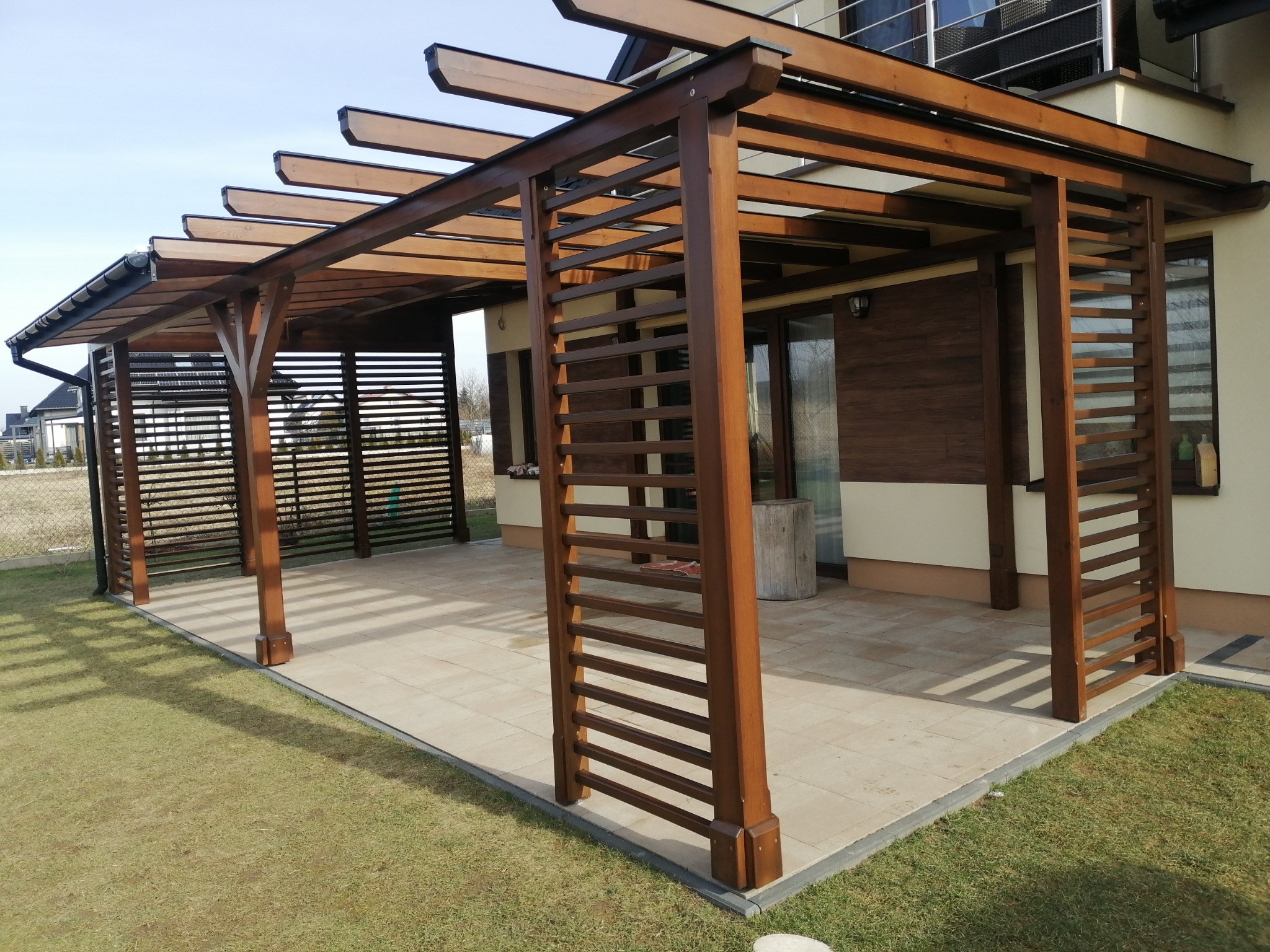 Zadaszona pergola drewniana z lamelami bocznymi, połączona z budynkiem, z widocznym trawnikiem i fragmentem dachu sąsiedniego domu.