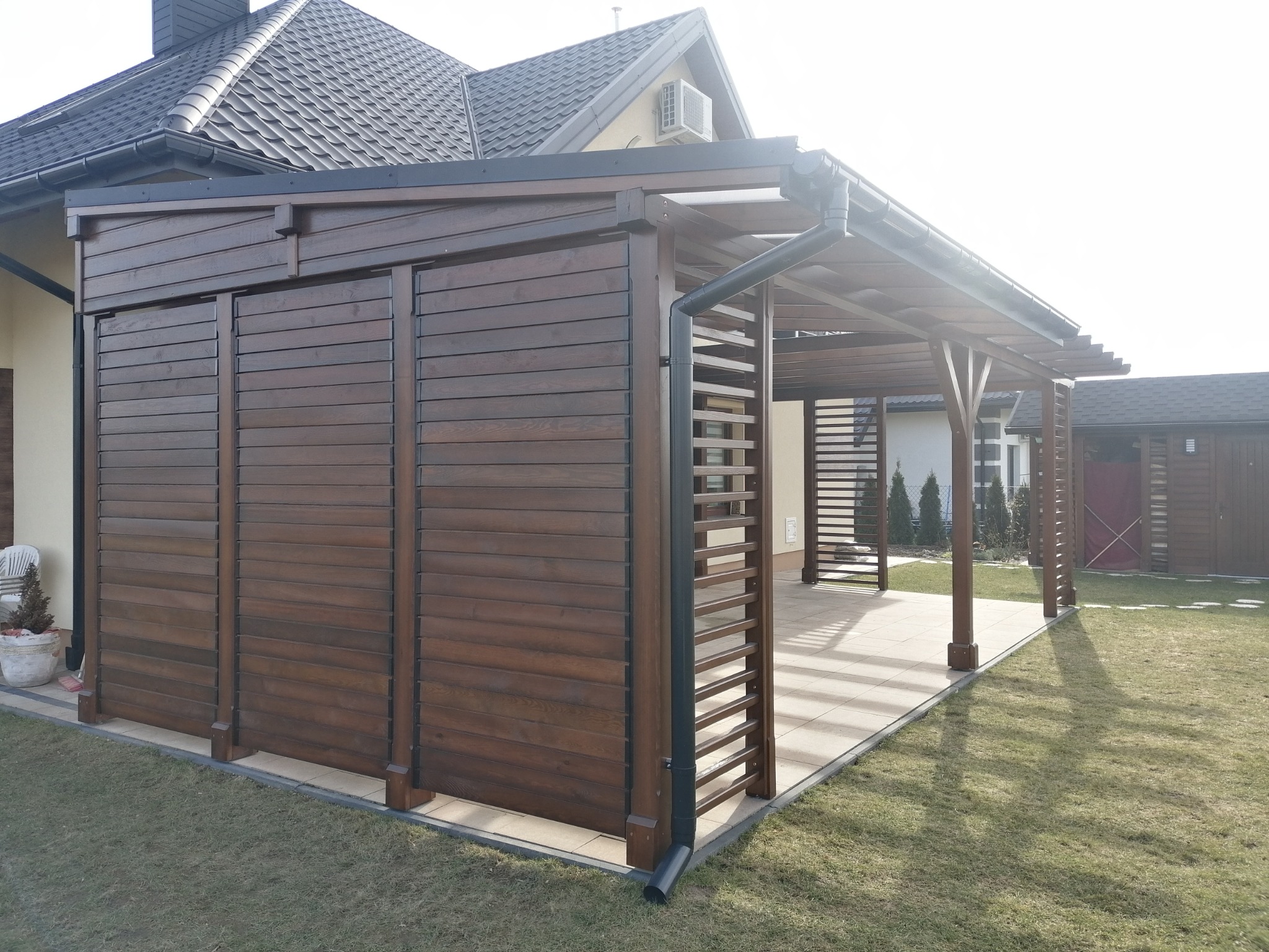 Zadaszona drewniana konstrukcja z poziomymi listwami, połączona z budynkiem, z widocznymi rynnami i fragmentem dachu domu w tle, na trawniku.