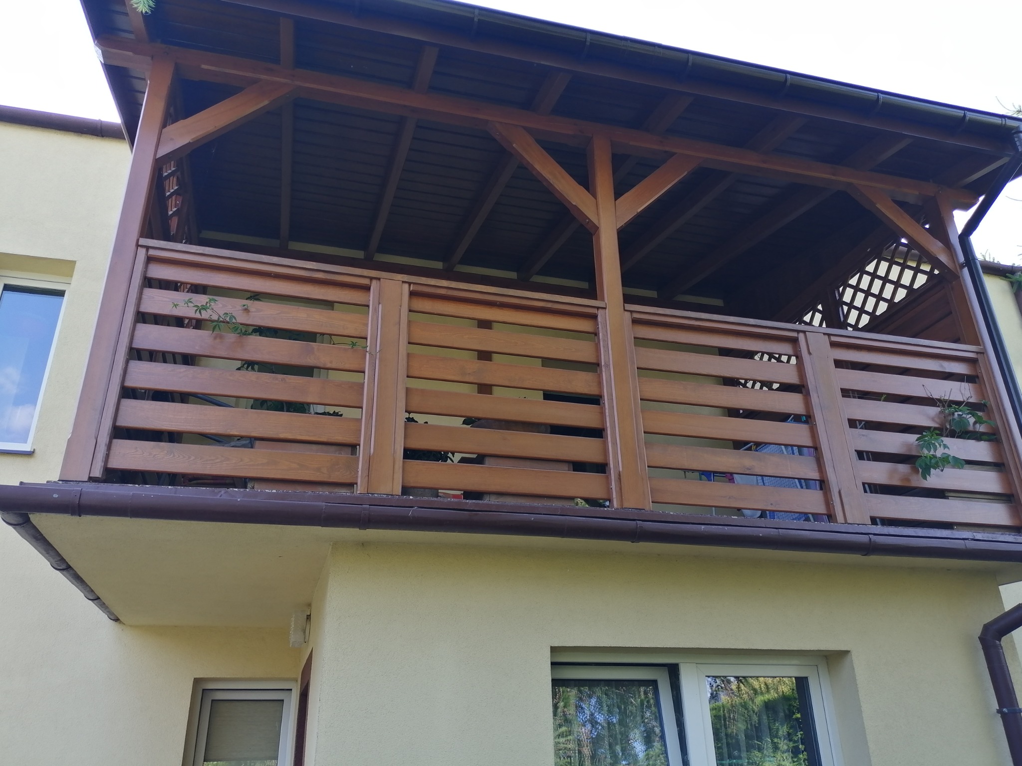 Zadaszony balkon z drewnianą balustradą z poziomych desek, widok z dołu, doniczkowe rośliny na balkonie, brązowe rynny.