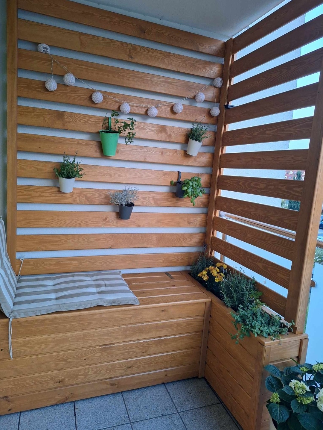 Drewniana zabudowa balkonu z siedziskiem i roślinami w doniczkach, oświetlona girlandą. Słoneczny dzień, widok z góry. Wyjątkowa aranżacja przestrzeni.