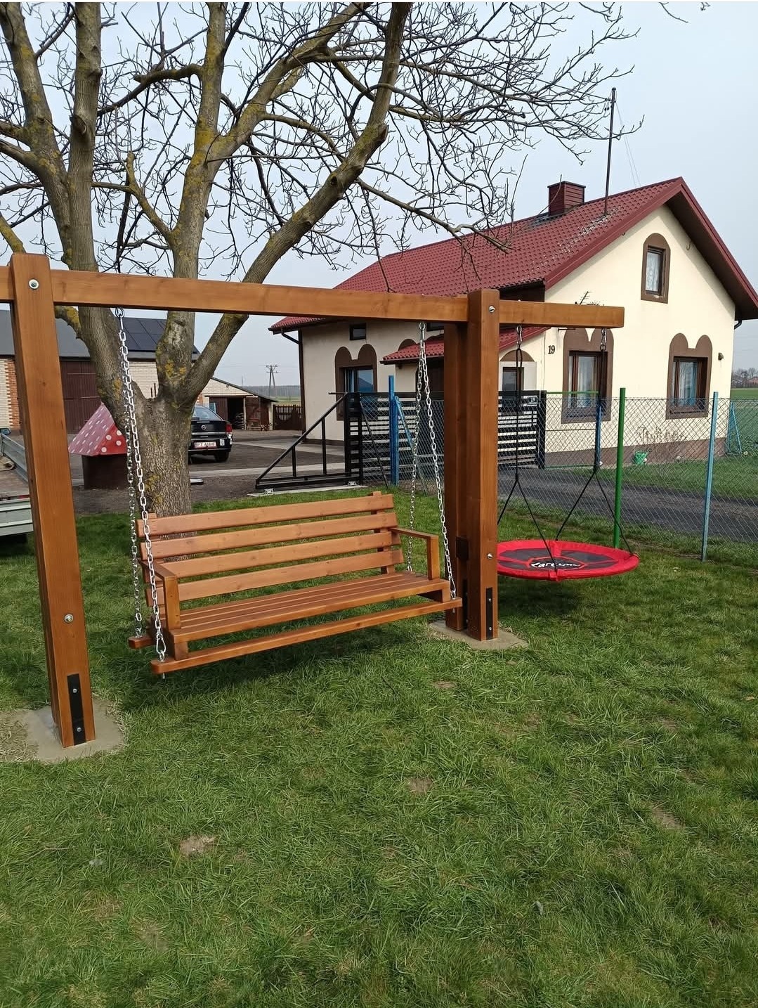 Drewniana huśtawka z siedziskiem i okrągłym elementem do huśtania, na tle domu z czerwonym dachem i zielonego trawnika. Widoczne mocowania huśtawki w ziemi.