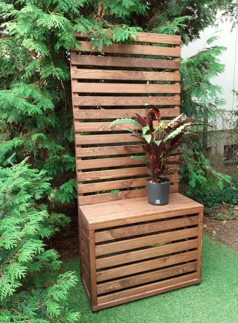 Drewniana konstrukcja z siedziskiem i wysokim ażurowym panelem, dekorowana rośliną w doniczce, na tle zieleni. Widoczne poziome deski i brązowe wykończenie.