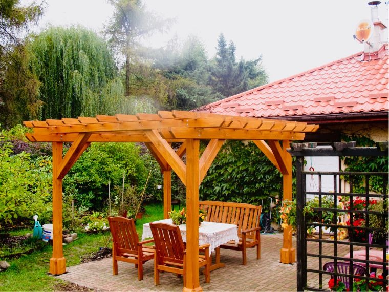 Drewniana konstrukcja zadaszenia nad stołem i krzesłami w ogrodzie, z widokiem na roślinność i fragment domu z czerwoną dachówką. Naturalne, ciepłe oświetlenie.