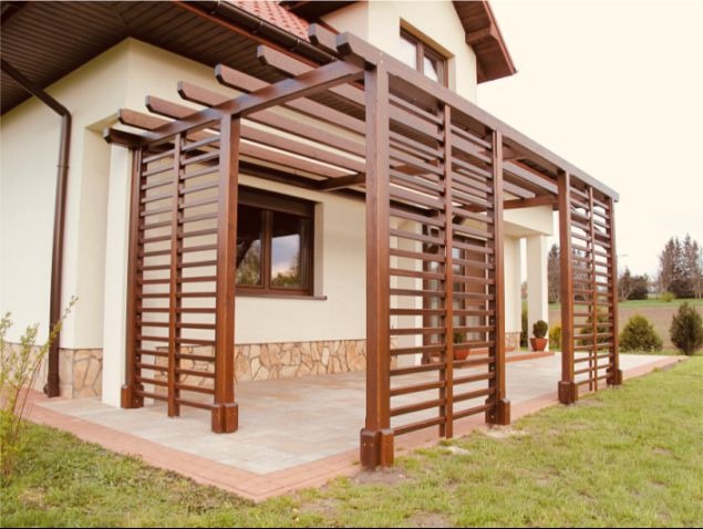 Elegancka, brązowa pergola z drewna, przylegająca do jasnego domu z kamienną elewacją. Widoczny fragment tarasu wyłożonego kostką i zadbany trawnik.
