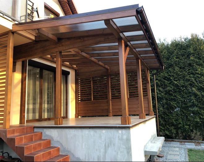 Nowoczesna, drewniana konstrukcja z zadaszeniem, prawdopodobnie taras lub weranda, z ażurowymi ściankami i schodami z płytek, na tle zieleni.