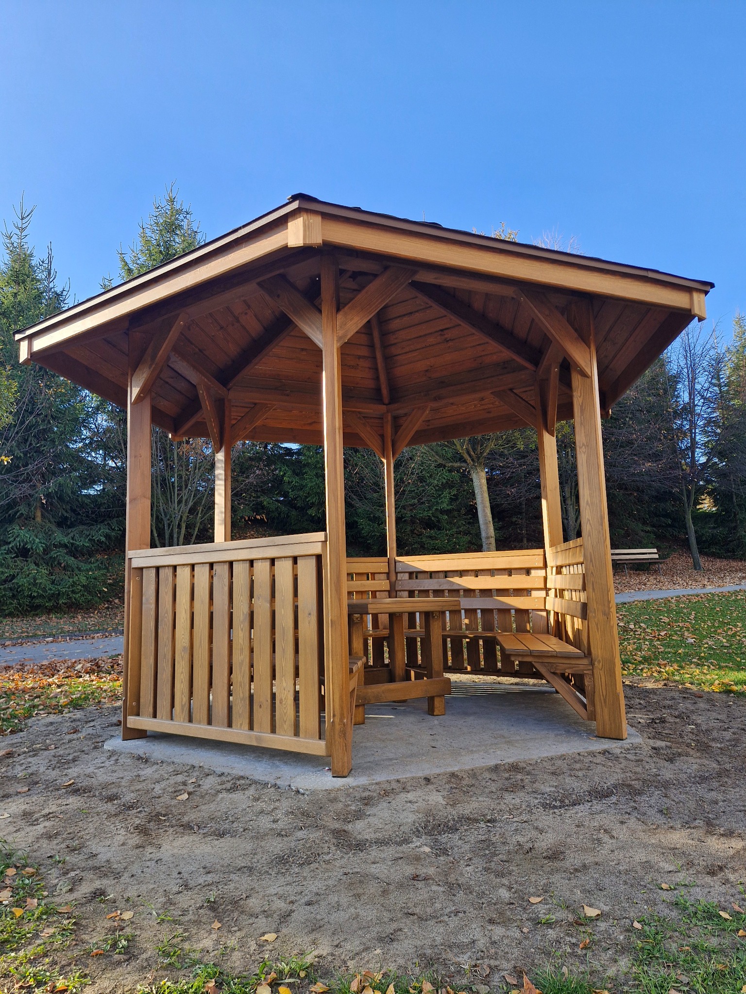 Drewniana altana z zadaszeniem i stołem z ławkami w parku, jesienne liście, iglaste drzewa w tle, słoneczny dzień, błękitne niebo.