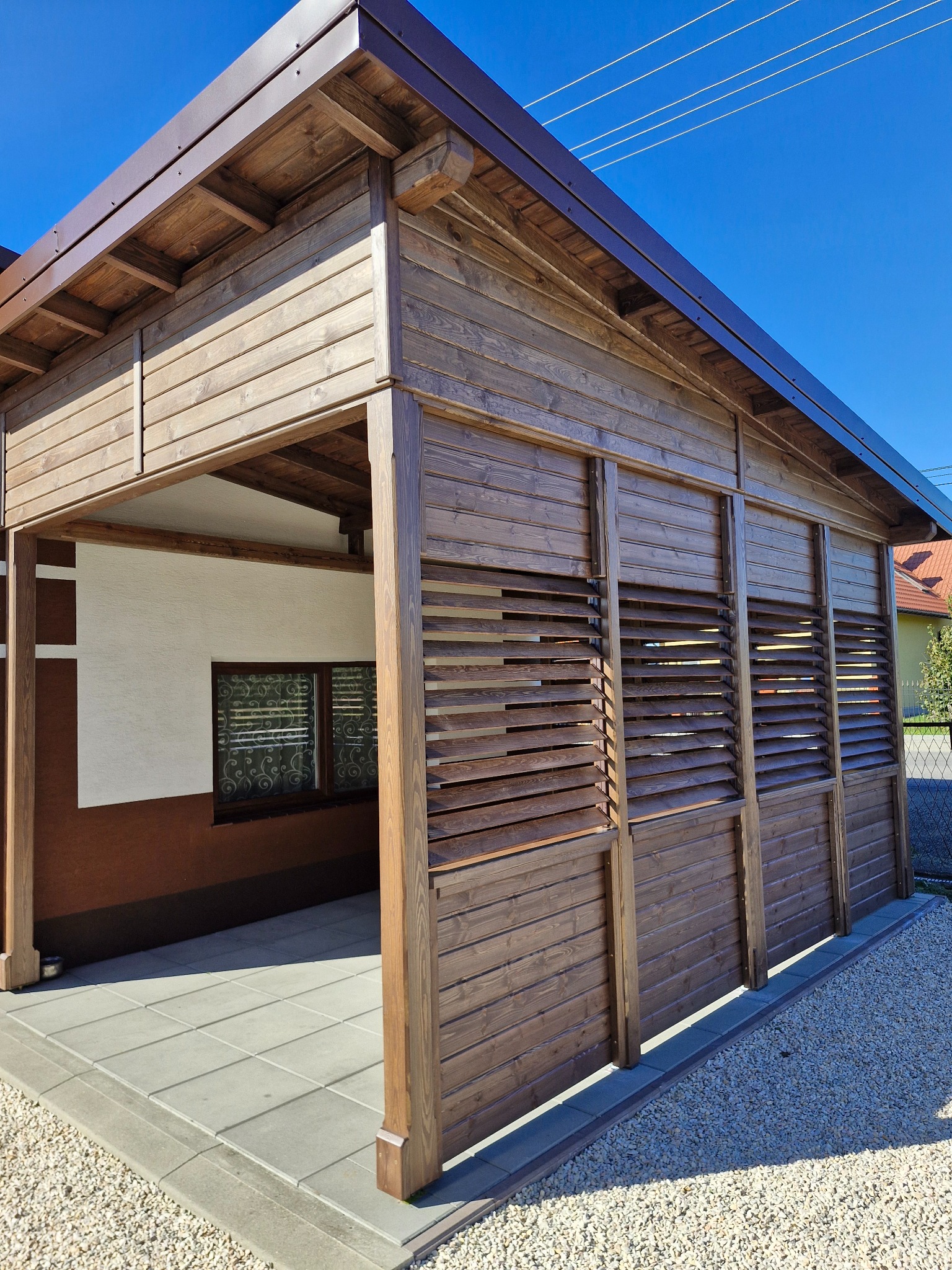 Zadaszona, drewniana konstrukcja z żaluzjowymi ścianami, prawdopodobnie altana lub pergola, z widoczną podłogą z szarych płyt i otoczeniem z jasnego żwiru.