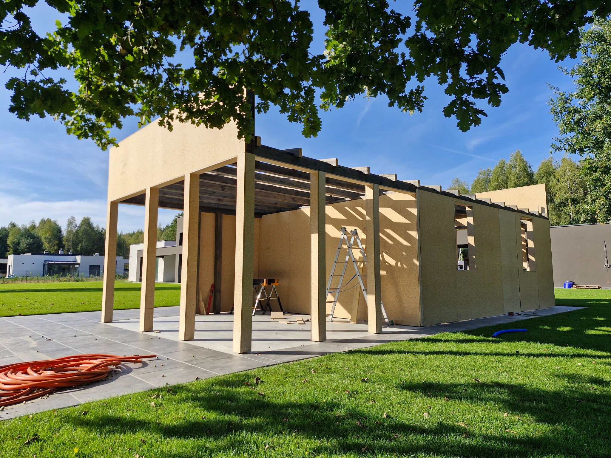 Konstrukcja z płyty OSB w ogrodzie, częściowo zadaszona, z widocznymi elementami szkieletu, drabina i narzędzia wewnątrz, trawnik i drzewa w tle.