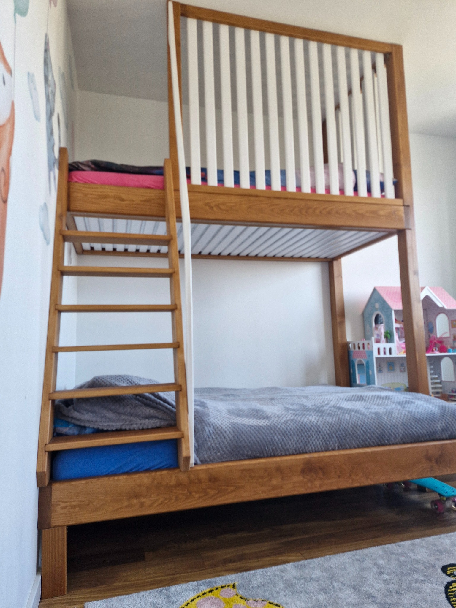 Drewniane łóżko piętrowe z drabinką, z białymi szczebelkami zabezpieczającymi górny poziom. W tle zabawki, na ścianie naklejki. Widok z dołu, na podłodze dywan.