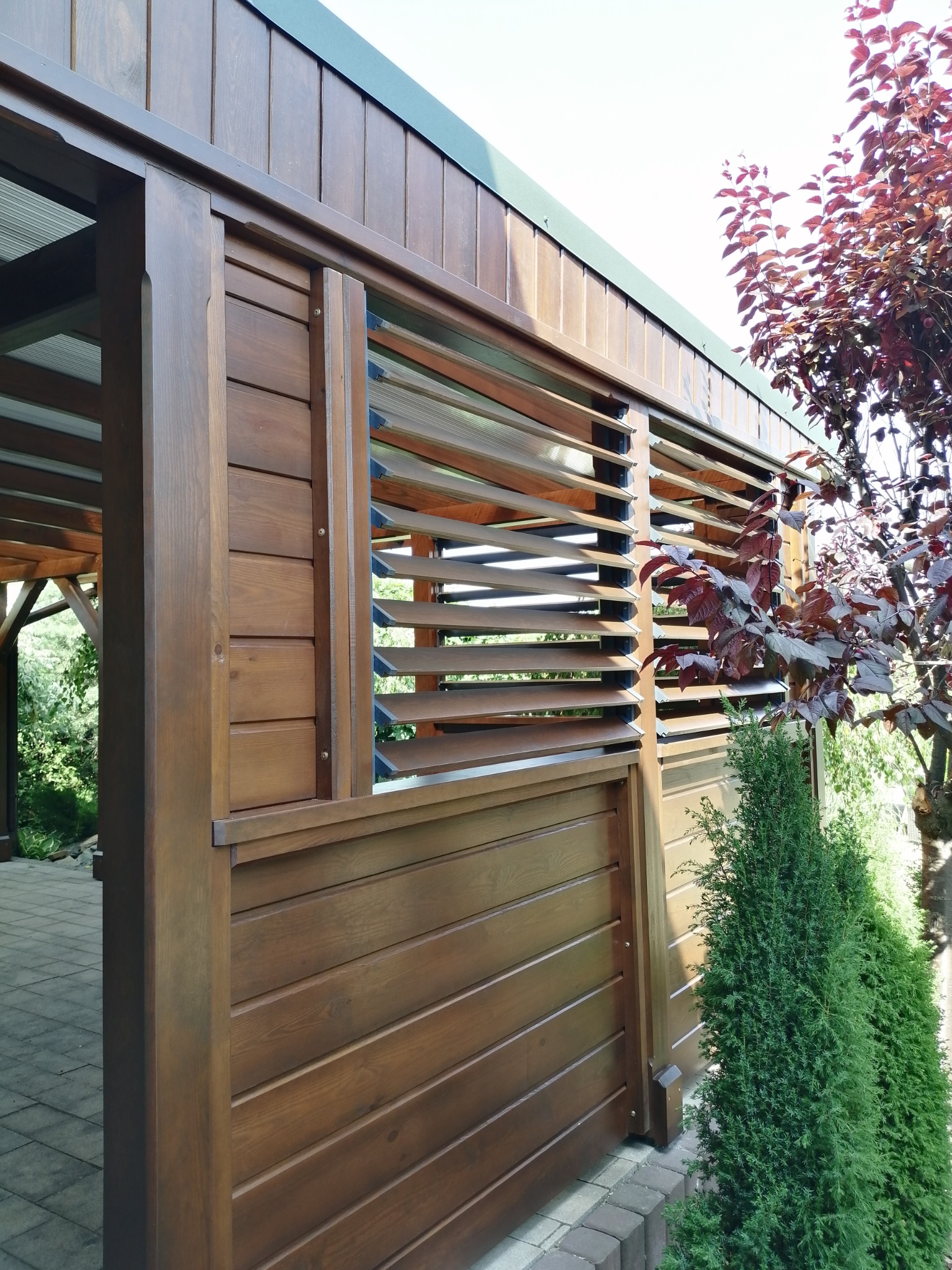 Drewniana konstrukcja z żaluzjami, prawdopodobnie altana lub pergola, wykończona w ciepłym odcieniu brązu, z widocznymi elementami roślinności w tle.