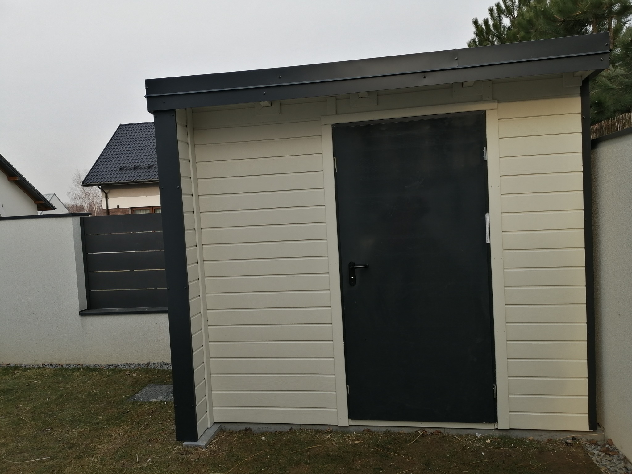 Nowoczesna, beżowa drewutnia z czarnymi elementami wykończeniowymi i drzwiami, na tle zielonego trawnika i ogrodzenia. Minimalistyczny styl, widoczne detale konstrukcyjne.