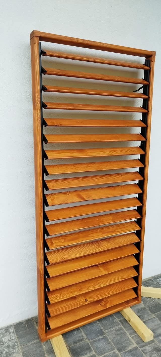 Drewniana konstrukcja z lamelami w odcieniach brązu, ustawiona pionowo, z widoczną strukturą drewna, umieszczona na drewnianych podporach.