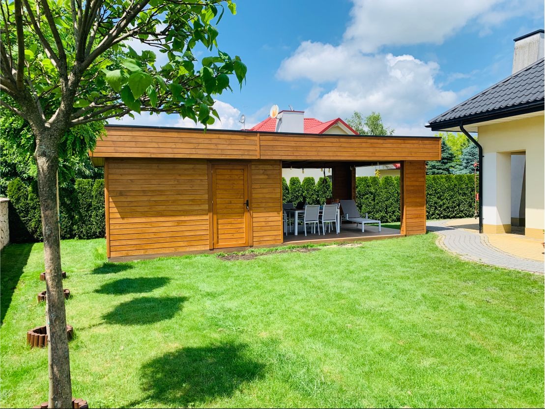 Nowoczesna, drewniana altana z zamykaną częścią i otwartą przestrzenią wypoczynkową z meblami ogrodowymi, otoczona zielonym trawnikiem i tujami.