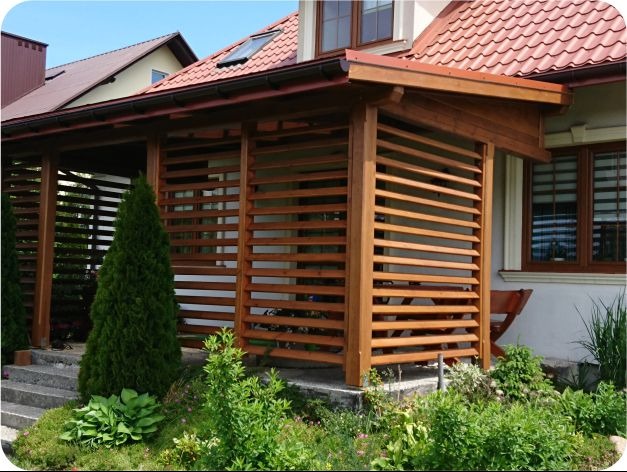 Drewniana pergola z poziomymi listwami, przylegająca do domu z czerwonym dachem. Widoczne rośliny i schody prowadzące do konstrukcji.