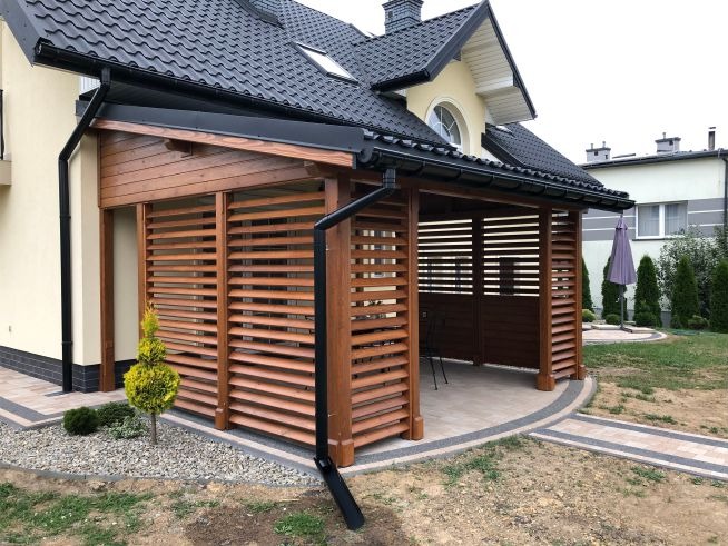 Drewniana konstrukcja z poziomymi listwami przy domu, z czarną rynną. Widoczny fragment ogrodu i kostka brukowa. Ujęcie z niskiej perspektywy.