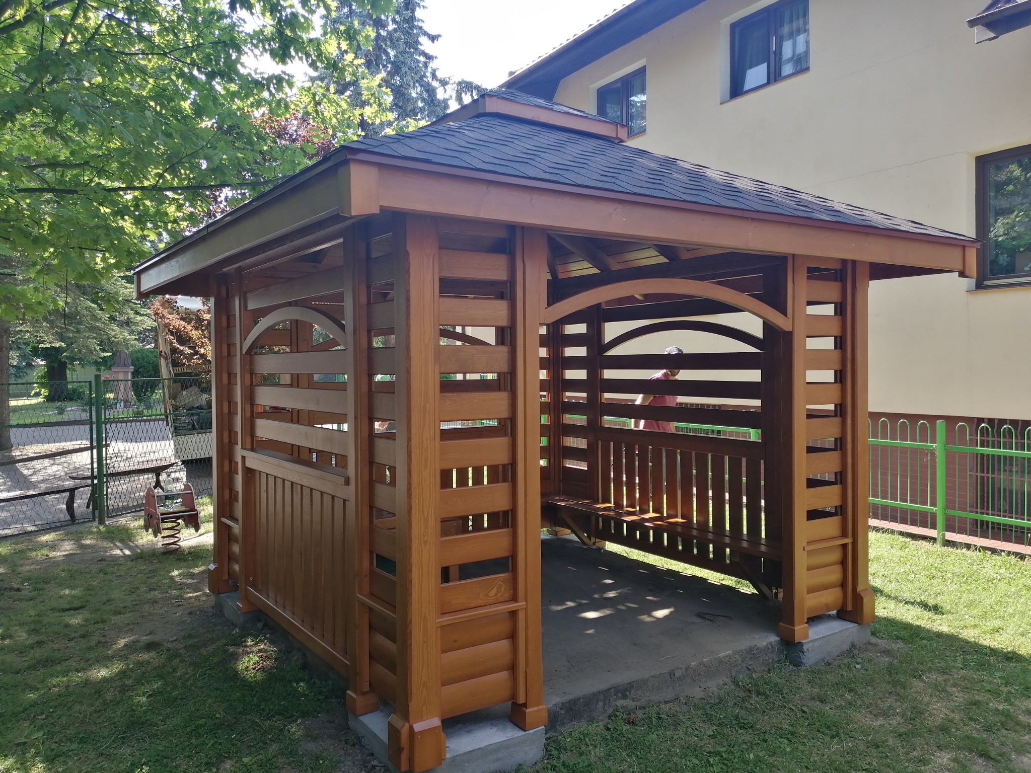 Drewniana altana ogrodowa z ławką wewnątrz, wykończona gontem bitumicznym, na betonowej podstawie, otoczona zielenią i budynkiem w tle.