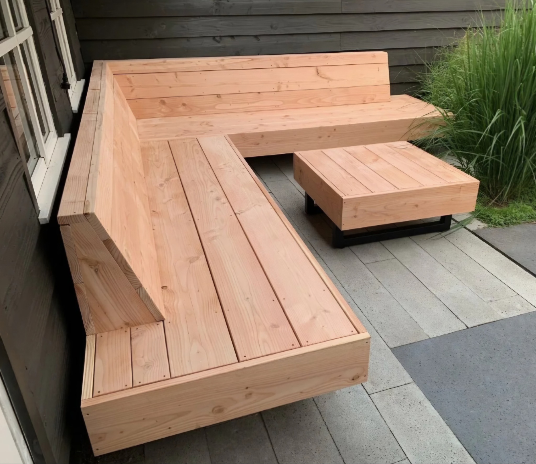 Drewniana, narożna ławka z niskim stolikiem na tarasie z szarych płyt. Prosta konstrukcja z jasnego drewna, w tle ściana budynku i wysoka trawa.