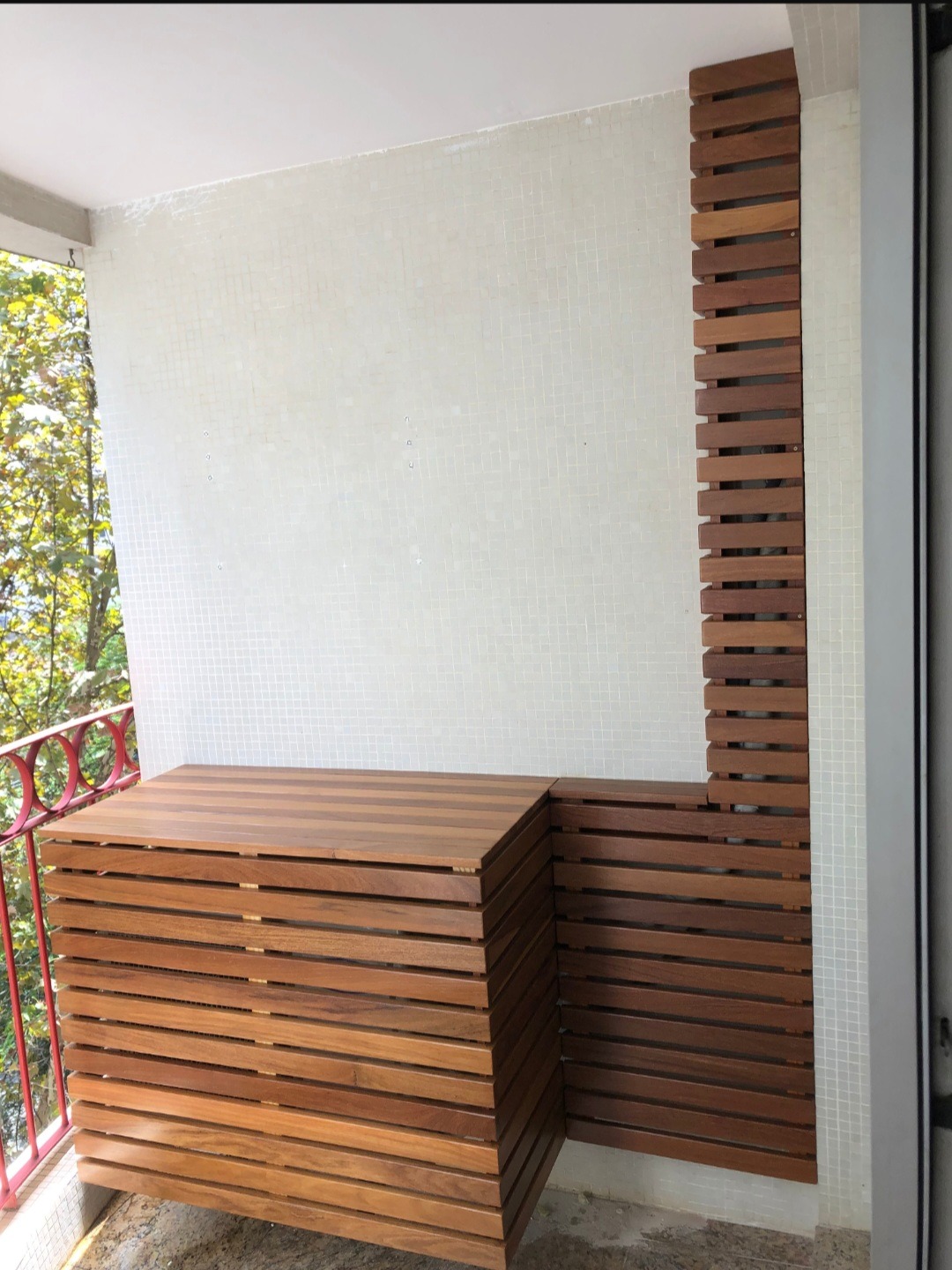 Zabudowa balkonu z drewna egzotycznego w formie skrzyni z poziomych listew i pionowej ścianki, widok z boku, balustrada balkonu z czerwonymi elementami.