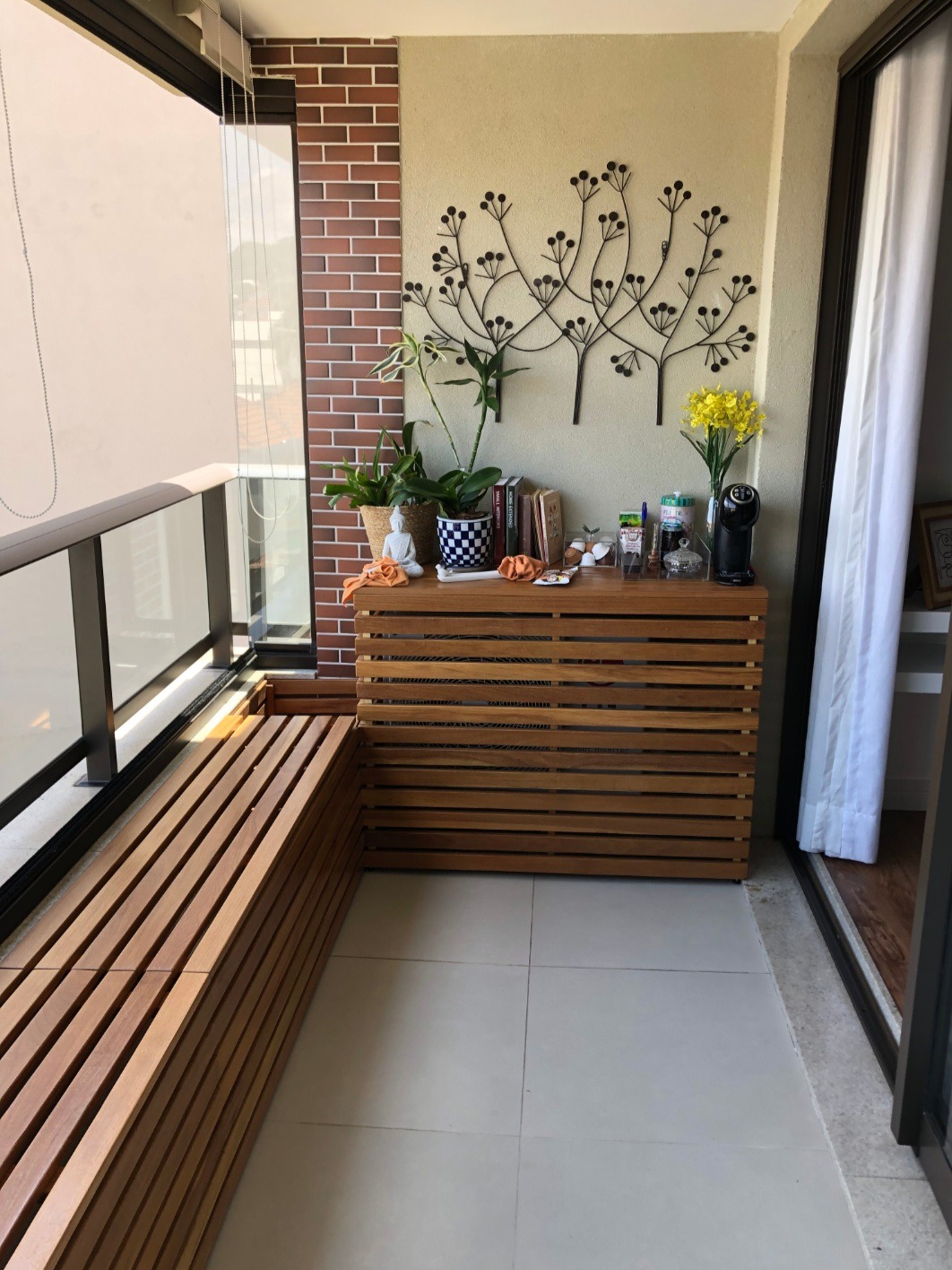 Funkcjonalny balkon z drewnianą zabudową, siedziskiem i dekoracjami. Wyjątkowy detal: metalowa ozdoba ścienna w kształcie drzewa. Jasne płytki podłogowe.