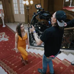 Kobieta w pomarańczowej sukience schodzi po schodach, filmowana profesjonalną kamerą stabilizowaną gimbalem, w tle bogato zdobione wnętrze z czerwonym dywanem i żyrandolem.