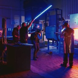 HOLE SP. Z O. O. - Ekipa filmowa kręci ujęcie w hucie szkła, oświetlając scenę niebieską lampą LED, z piecem hutniczym w tle i pracownikiem w stroju ochronnym.